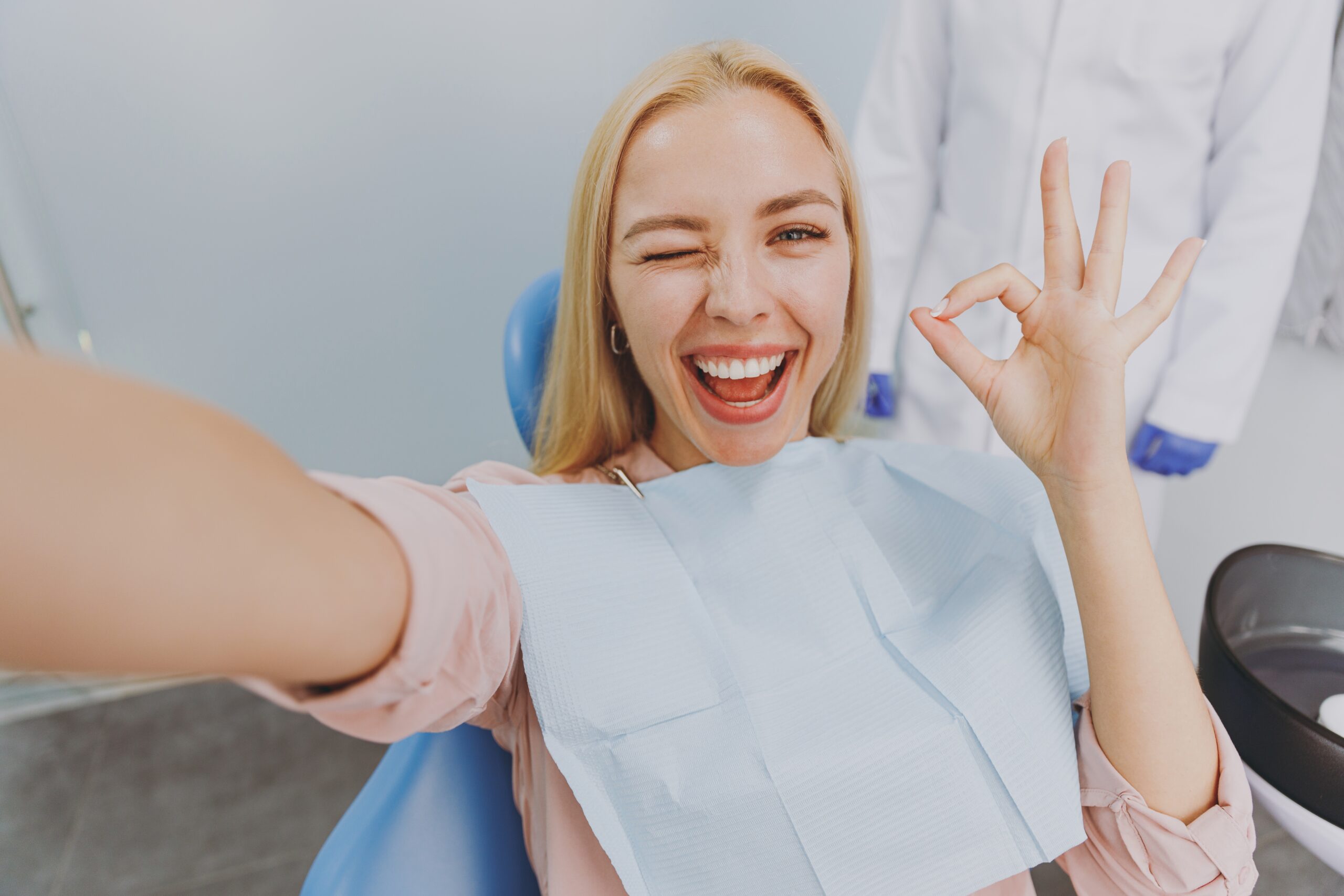 happy female dental patient taking a selfie and smiling at at the camera in a blue dental seat.