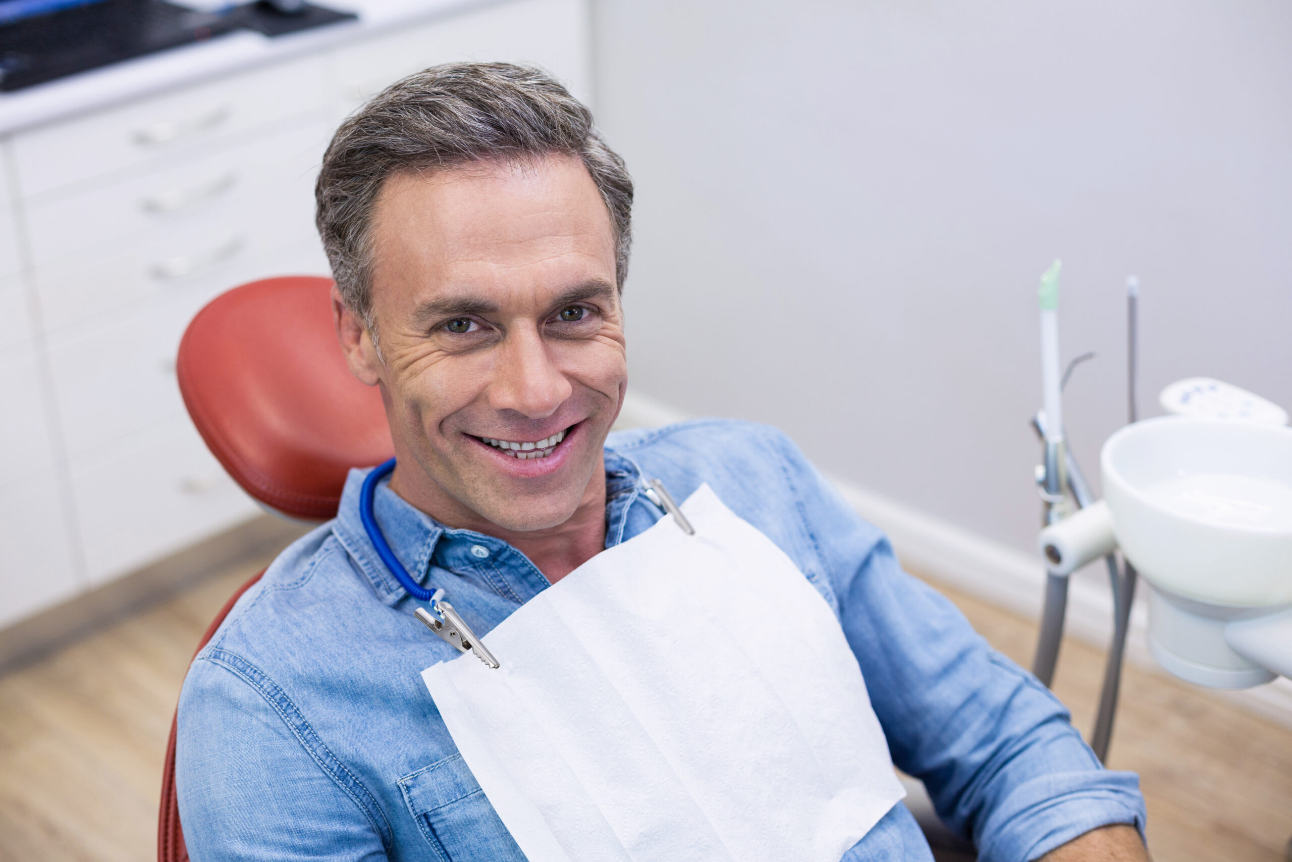 male dental patient wearing a blue button up shirt sitting in a red dental chair smiling at the camera